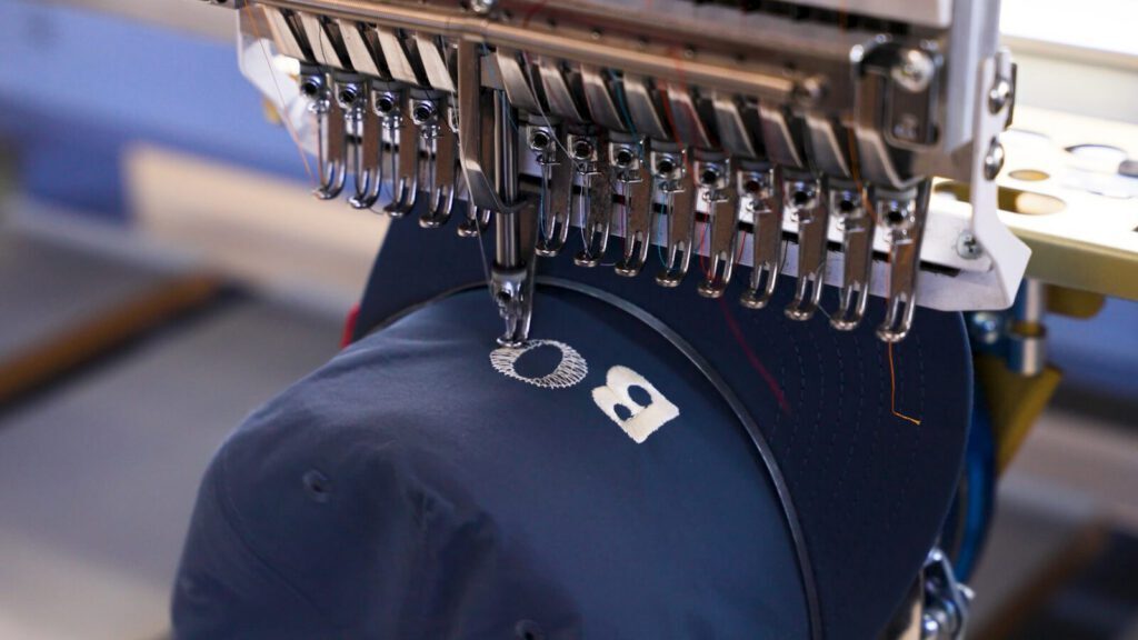 A row of multi-head Barudan embroidery machines stitching navy caps on Prodigy Promos' production floor in Lindon, Utah