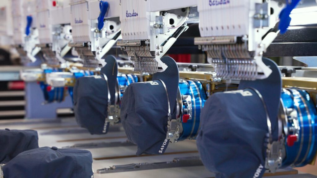 Looking down Prodigy Promos' embroidery line as Barudan heads stitch logos into navy caps held in framing hoops, Lindon, Utah