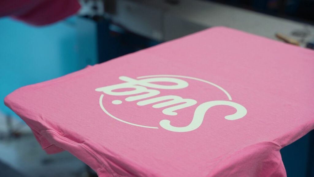 A pink Swig tee on the platen of a screen-printing press at Prodigy Promos in Lindon, Utah