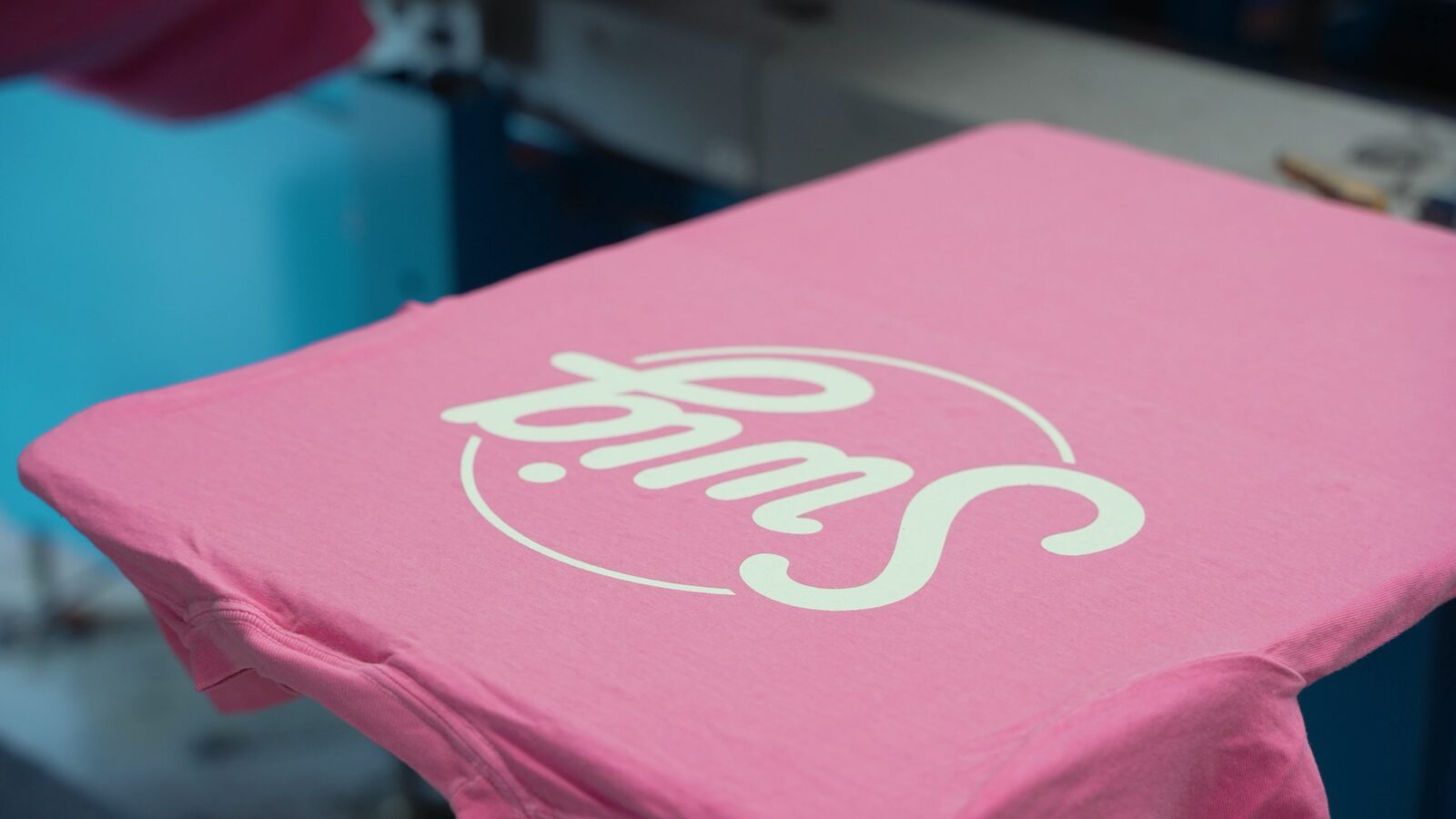 A pink Swig tee on the platen of a screen-printing press at Prodigy Promos in Lindon, Utah
