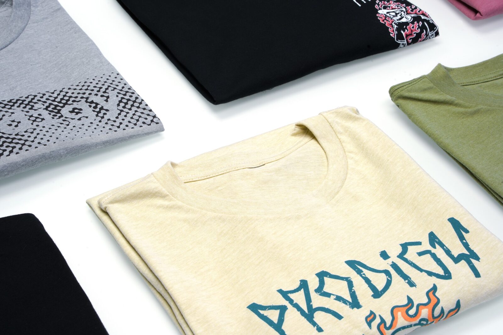 Neatly folded custom-printed t-shirts in a flat lay - gray halftone, black, cream, olive, and pink Prodigy sample tees
