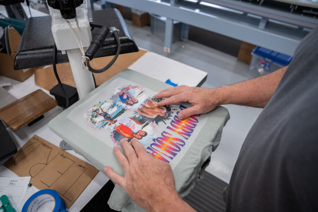 Hands positioning a full-color DTF transfer onto a sage green tee at the heat press