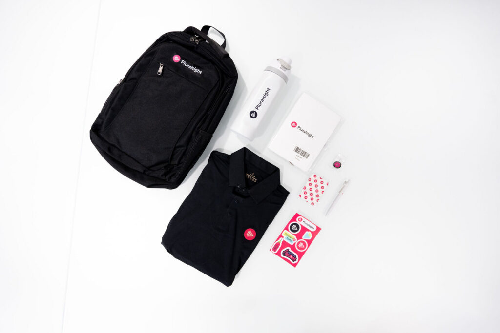 Pluralsight onboarding kit laid out: backpack, polo, tee, water bottle, notebook and sticker pack