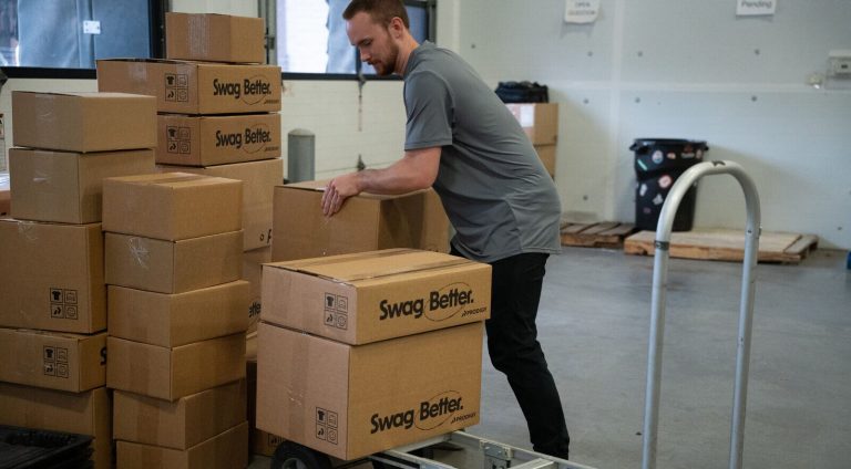 A Prodigy Promos team member loading branded Swag Better shipping cartons onto a hand truck in the Lindon fulfillment area.