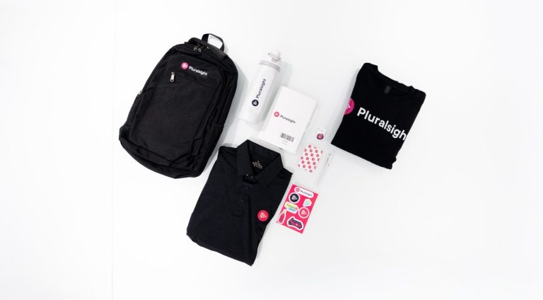 A Pluralsight new-hire welcome kit laid flat: black backpack, white water bottle, notebook, black polo, black tee, pen, pin, and a pink sticker sheet.