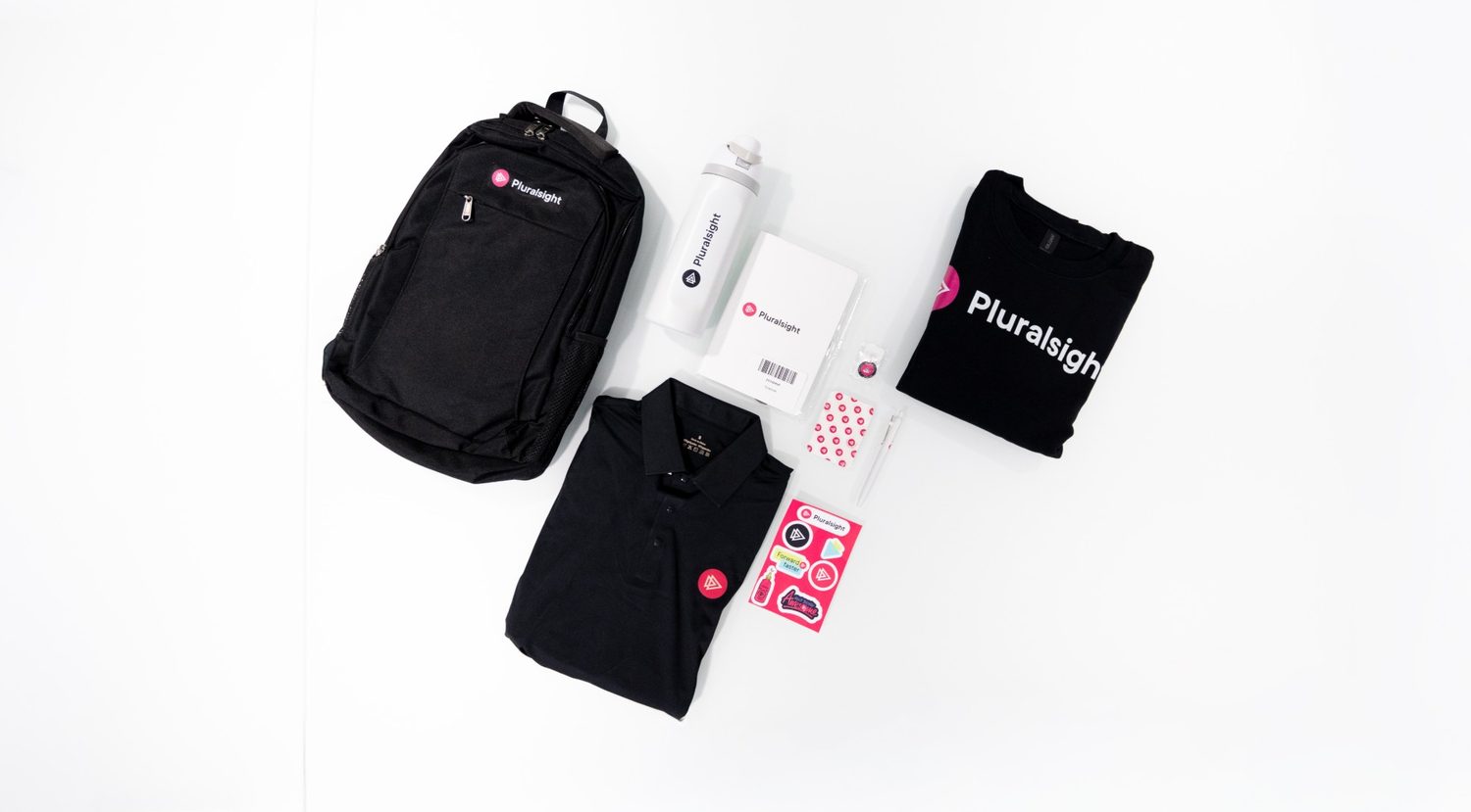 A Pluralsight new-hire welcome kit laid flat: black backpack, white water bottle, notebook, black polo, black tee, pen, pin, and a pink sticker sheet.